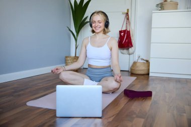 Laptop ve kablosuz kulaklık kullanan genç ve rahat kadın portresi, meditasyon yaparken evde lastik minder üzerinde yoga yapmak, Asya 'da oturmak..