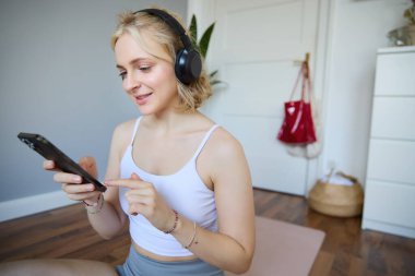 Atletik genç kadın portresi, cep telefonu tutmak ve kablosuz kulaklık takmak, evde egzersiz, spor uygulaması kullanmak.