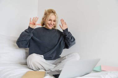Yatağında çalışan genç gülümseyen bir kadının portresi, yatak odasında dizüstü bilgisayar ve belgelerle oturuyor, video sohbeti sırasında el sallıyor, özel öğretmen öğrencisini internet üzerinden selamlıyor.