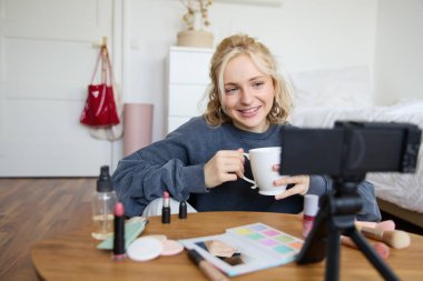 Blogculuk, modern teknoloji, e-ticaret ve insan konsepti. Mutlu gülümseyen kadın video blogcusu elinde çay bardağıyla evde video blogu için güzellik dersi kaydederken makyaj yapıyor..