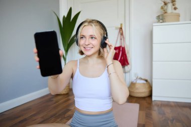 Formda ve sağlıklı kadın egzersizlerinin portresi, kulaklık takması, akıllı telefon ekranı göstermesi, boş siyah ekran. Refah ve yaşam tarzı kavramı