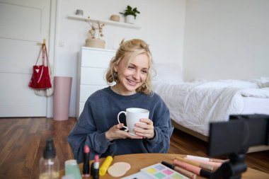 Güzel sosyal medya güzellik blogcusu portresi. Yatak odasında, dijital kameranın önünde oturmuş çay içiyor, sohbet ediyor, takipçilerle konuşuyor..