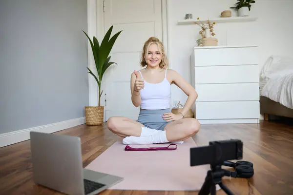 Fitness blogger, woman records video of herself, sitting on rubber mat for exercises in front of digital camera and laptop, shows thumbs up, creates content for social media account about wellbeing.