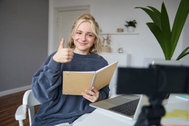 Gülümseyen, pozitif genç bir kadın, başparmak kaldırıyor, dijital kamera ve dizüstü bilgisayarla aynı odada oturuyor, video kaydediyor, online ders veriyor, takipçiler için içerik oluşturuyor..