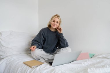 Evde çalışan genç bir kadının portresi, defterler, belgeler ve dizüstü bilgisayarlarla dolu bir yatakta oturuyor, mutlu görünüyor ve kameraya gülümsüyor..