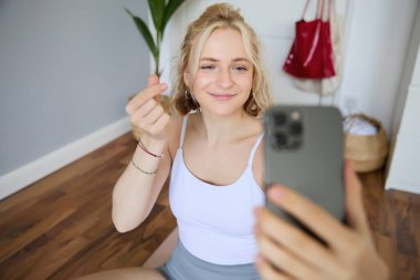 Genç, güzel sarışın bir kadının portresi, evde yoga minderi üzerinde egzersiz yapan beden eğitimi öğretmeni, akıllı telefondan selfie çekmesi, sosyal medya hesabına spor hakkında kendi videosunu kaydetmesi..