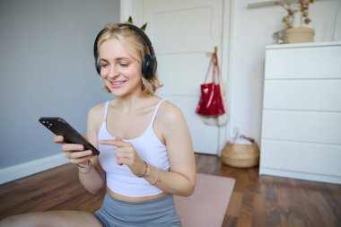 Kablosuz kulaklıklı genç bir kadının portresi, akıllı telefonuyla lastik minderin üzerinde oturuyor, egzersiz uygulaması kullanıyor, telefonunda fitness uygulaması kullanıyor, kulaklıkla dinliyor..