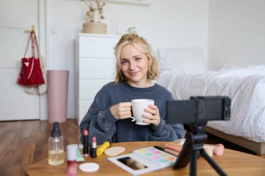 Güzel sosyal medya güzellik blogcusu portresi. Yatak odasında, dijital kameranın önünde oturmuş çay içiyor, sohbet ediyor, takipçilerle konuşuyor..