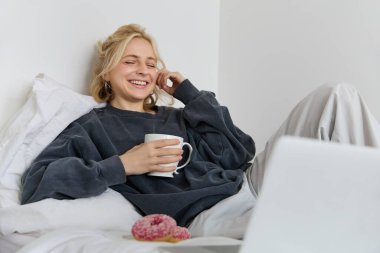 Mutlu kadın portresi evde tatilden zevk alıyor, yatakta uzanıyor, dizüstü bilgisayarda televizyon seyrediyor, ekrana bakıyor ve gülümsüyor, çay içiyor ve yatak odasında yemek yiyor..