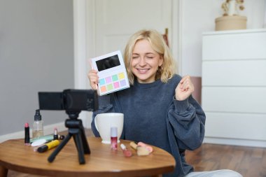 Heyecanlı gülümseyen sarışın kadın yeni bir göz falı paletinde video eleştirisi kaydediyor, takipçilere ürün gösteren yaşam tarzı blogcusu, coşkulu bir yüzle yerde oturuyor..