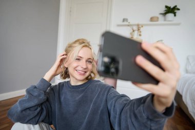 Genç bir kadının portresi, sosyal medya etkisinde, odasında selfie çekiyor, yerde oturuyor, akıllı telefon tutuyor ve fotoğraf için poz veriyor..