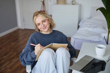 Gülümseyen tatlı bir kadının portresi, yaşam tarzı blogcusu, günlük günlük günlük veya planlamacıyla odasında oturuyor, dijital kameraya video kaydediyor, sosyal medya için günlük rutin hakkında içerik oluşturuyor.