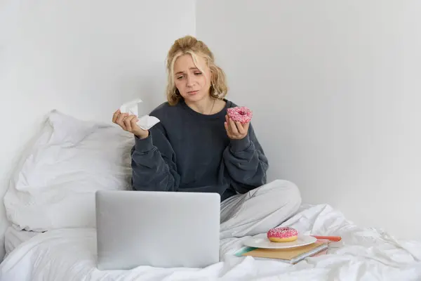 Portrait of sad, crying young woman, staying at home, sitting in bed with doughnut and comfort food, looking at something upsetting on laptop screen.