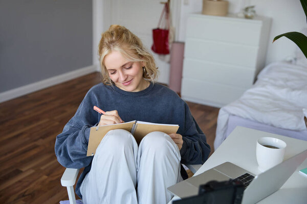 Portrait of young woman writing in diary, making notes in notebook, sitting on chair in a room in front of laptop, student doing homework.