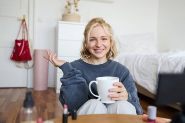 Neşeli kadın, güzellik blogcusu, dijital kamerada yaşam tarzı vlog 'unu kaydediyor, gündelik konuşuyor, sosyal medya takipçileri için bir hikaye anlatıyor, fincanı tutuyor, çay içiyor ve odasında yerde oturuyor..
