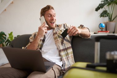 Kredi kartı tutan ve dizüstü bilgisayar kullanan bir adamın resmi. Tatil için bavul ve kamera hazırlamış. Turizm konsepti