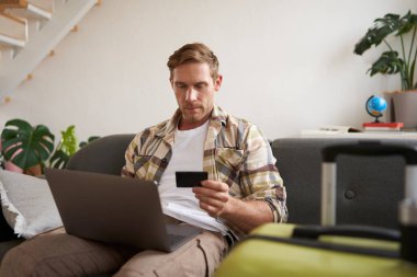 Neşeli bir adamın portresi, tatile çıkıyor, yurtdışına seyahat planları yapıyor, dizüstü bilgisayarı açık, oturma odasında bavuluyla oturuyor, bir otel rezervasyonu yaptırıyor..