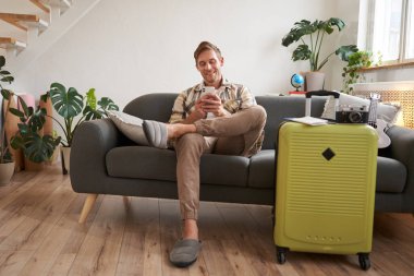 Yakışıklı genç adam kanepede bavuluyla oturuyor, elinde akıllı telefon, kitap pansiyonu ya da tatil tatili var, cep telefonuyla sohbet ediyor. Turizm ve seyahat kavramı