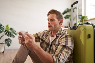 Bavullu bir adam portresi, yere oturmuş, cep telefonuyla oynuyor, ciddi görünüyor. Turizm ve halk konsepti