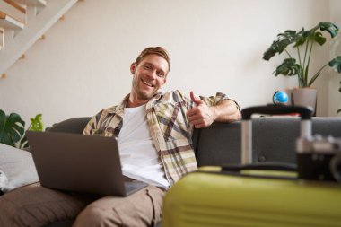 Dizüstü bilgisayarı olan neşeli bir adam, turist valizin yanına oturur, başparmağını kaldırır, tatil için yer ayırtır, tatile gider. Turizm konsepti