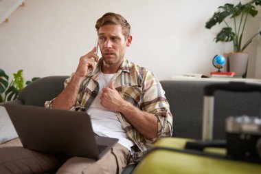 Kafası karışmış bir adam, cep telefonuyla konuşuyor, dizüstü bilgisayarla oturuyor, yanına bavulu yerleştiriyor, seyahat acentasıyla tatille ilgili sorular soruyor..