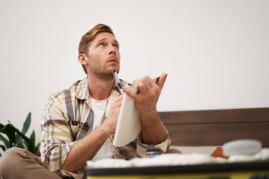 Bavullu bir adamın portresi, tatil için otururken ve eşyalarını toplarken, tatil için eşya listesini kontrol ederken, not alırken, not defterine not alırken. Yaşam tarzı ve turizm konsepti