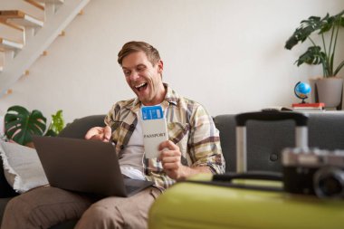 Hevesli yakışıklı bir adam, iki uçuş bileti ve pasaportu olan bir turist, dizüstü bilgisayara bakıyor ve tatilde zevkle bağırıyor. Turizm konsepti