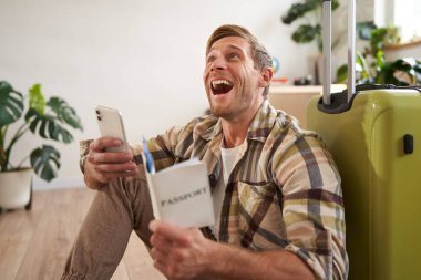 Pasaportu, biletleri ve akıllı telefonu olan mutlu genç bir adam, gülüyor, bavuluyla oturuyor, tatile çıkıyor. Halk ve turizm kavramı