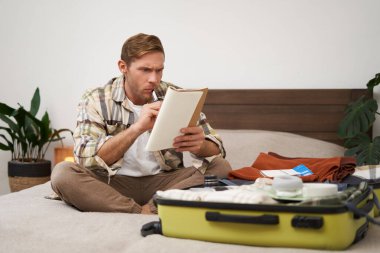 Kızgın bir adamın portresi, bavul hazırlıyor, tatil için kontrol listesiyle defterde hayal kırıklığına uğramış görünüyor, tatile çıkmaya hazırlanıyor. Turist ve insan konsepti