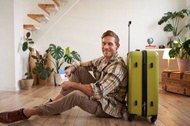 Gülümseyen yakışıklı adamın portresi, bavuluyla yerde oturuyor, elinde pasaport ve uçak biletleri var, turneye çıkıyor..