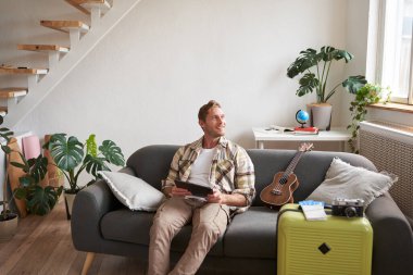 Yakışıklı genç adamın portresi, bavul ve iki bilet yanında dijital tabletle oturan turist, tatili için internetten bir şeyler ayırtıyor, tatil hazırlıyor..