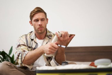 Elbiseleriyle valizine ciddi şekilde bakan genç bir adamın portresi, kontrol listesini gözden geçiriyor, elinde notlarla defteri tutuyor, tatilde ne getireceğini seçiyor. Turizm konsepti.