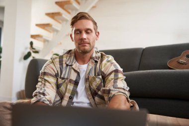 Bilgisayarı kullanan genç adamın portresini kapat, bilgisayara bak, yerde cihazıyla otur, web sitelerine bak.