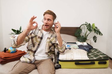 Yakışıklı sarışın adamın portresi, turist tatile çıkıyor, kablosuz kulaklıkla müzik dinliyor, kıyafetlerini bavula koyuyor, tatil yerini seçiyor, iyi el hareketi gösteriyor..