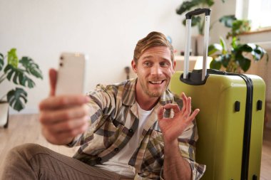 Bavullu, gülümseyen yakışıklı bir adamın portresi, akıllı telefondan selfie çekiyor, tamam işareti gösteriyor, tatil yerini onaylıyor, sosyal medya hesabına fotoğraflar yüklüyor..