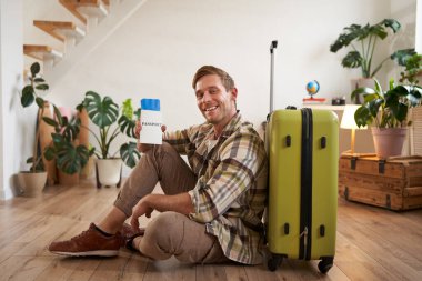 Gülümseyen yakışıklı adam pasaportuna ve uçuş biletine bakar, bavuluyla yere oturur..