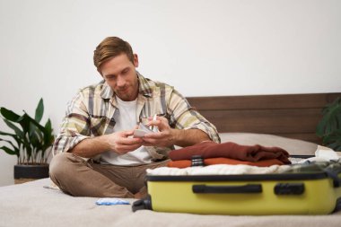 Bavullu genç bir adamın portresi, uçak biletlerini elinde tutması, tatile gitmeye hazırlanması, tatil için eşyalarını toplaması, turist kıyafetlerini çantaya koyuyor..