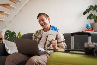 Mutlu turist portresi, dizüstü bilgisayara heyecanlı bakan bir adam, uçak biletleri tutuyor, tatile gidiyor, tatil için heyecanlı, bavuluyla oturma odasında oturuyor..