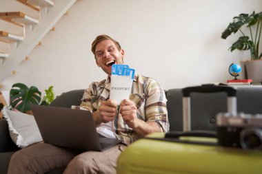 Mutlu yakışıklı adamın portresi, uçuş biletlerini gösteriyor, yaz tatili için yer ayırtıyor, dizüstü bilgisayarla oturma odasında oturuyor, kameraya gülümsüyor..