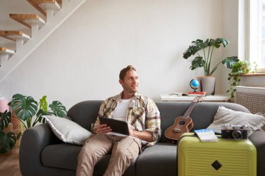 Tabletli genç bir adamın resmi, oturma odasında bavuluyla oturuyor, okuyor, rotayı okuyor, tatile hazırlanıyor. Turizm konsepti