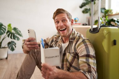 Elinde bavuluyla gülümseyen yakışıklı bir adamın portresi, akıllı telefona bakıyor, seyahat bileti ve pasaport tutuyor. Turizm ve seyahat kavramı