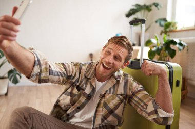 Gülümseyen yakışıklı adamın portresi fotoğrafta bisepslerini gösteriyor, akıllı telefonda selfie çekiyor, bavulun yanına oturuyor, tatil turuna çıkıyor..