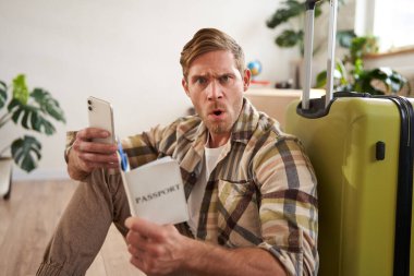 Adam kafası karışmış görünüyor, akıllı telefon kullanıyor, pasaport tutuyor, bagajın yanında oturuyor, seyahatini ya da tatilini planlıyor. Boşluğu kopyala