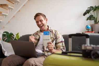 Heyecanlı genç adam, dizüstü bilgisayarında mutlu görünüyor, uçuş biletleri ve pasaportuyla oturuyor, tatil gezisi için bavulunu hazırlıyor..