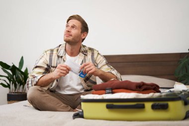 Bavullu genç bir adamın portresi, uçak biletlerini elinde tutması, tatile gitmeye hazırlanması, tatil için eşyalarını toplaması, turist kıyafetlerini çantaya koyuyor..