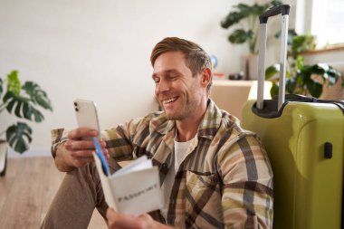 Gülümseyen turist portresi, bavulla oturan, elinde akıllı telefon, pasaport ve biletler olan adam tatile çıkıyor. Turizm ve halk konsepti