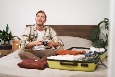 Heyecanlı genç bir adamın portresi, bavuluyla yatakta oturuyor, tatile çıkıyor, dijital kamerayla fotoğraf albümünü inceliyor, yaz tatiline hazırlanıyor..