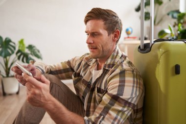 Bavullu bir adam portresi, yere oturmuş, cep telefonuyla oynuyor, ciddi görünüyor. Turizm ve halk konsepti