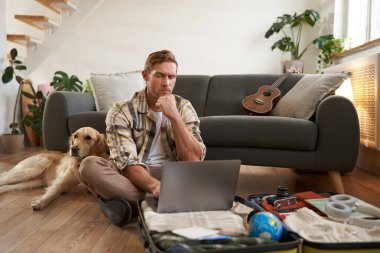 Genç bir adamın resmi, tatile çıkan bir turist, oturma odasında bir köpekle oturuyor, dizüstü bilgisayara bakıyor, bilgisayardaki bir uçağa giriş yapıyor. Turizm ve seyahat kavramı