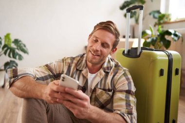 Sosyal medyada gülümseyen yakışıklı bir adamın portresi, akıllı telefondaki mesajla gülüşmeler, mutlu yüzlü cep telefonuna bakmalar, yerde valizin yanında oturmalar..